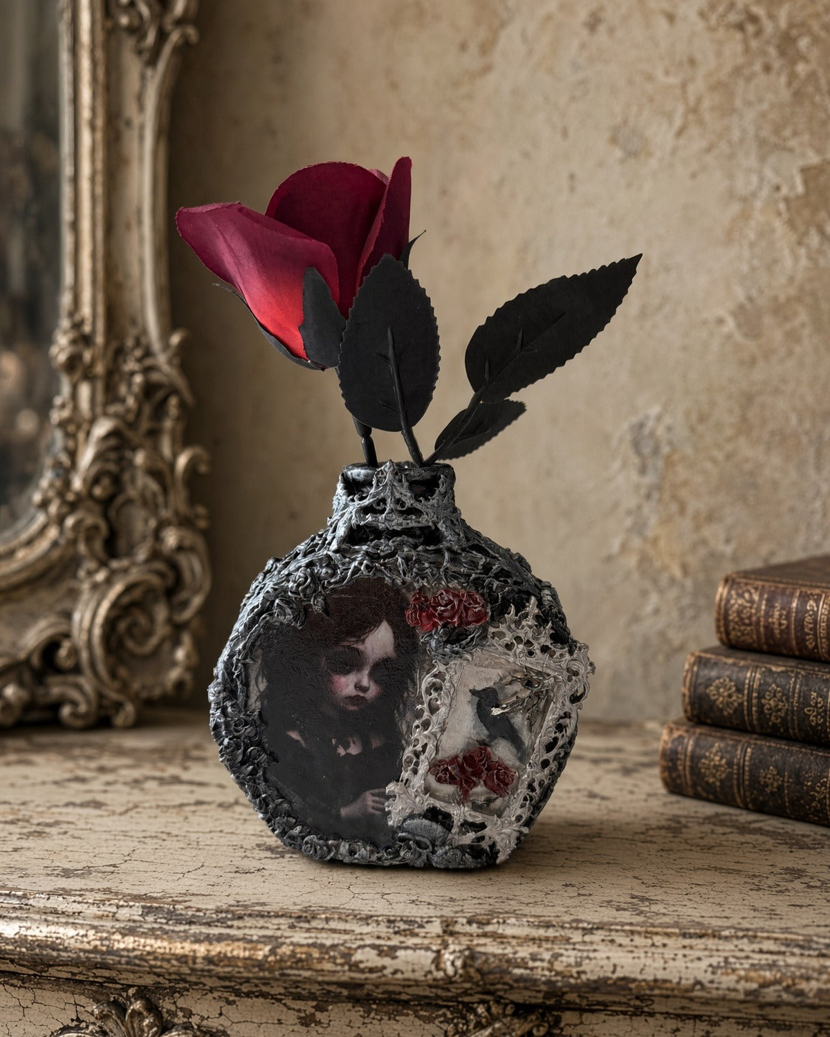 Handmade gothic decoupage glass bottle with doll-like girl, crow in silver frame and red roses, styled with a red rose, on cream table